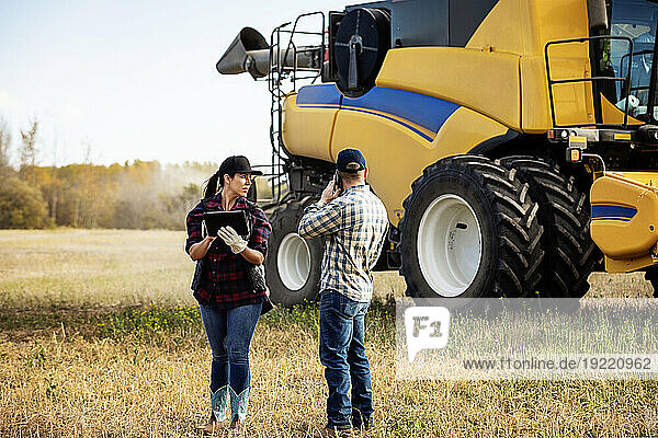 Husband and wife standing next to a combine harvester and using portable wireless devices to manage and monitor their fall  canola harvest yield; Alcomdale  Alberta  Canada