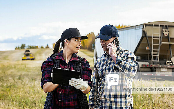 Close-up of a husband and wife using portable wireless devices to manage and monitor their canola harvest  making a call with a tractor trailer hauler and combine harvester in the background; Alcomdale  Alberta  Canada