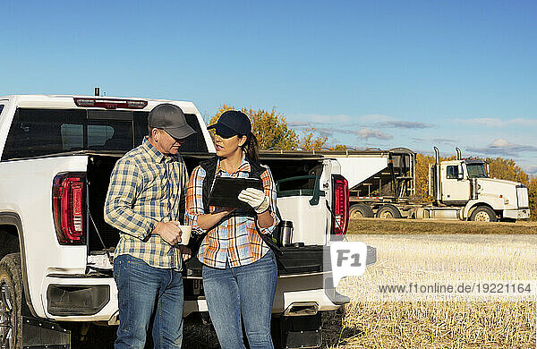 Husband and wife taking time for a coffee while leaning on the tailgate of their half-ton truck and using a portable wireless device to manage and monitor their canola harvest with a semi-trailer grain hauler in the background; Alcomdale  Alberta  Canada