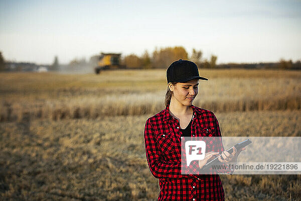 Portrait of a young farm woman standing in a grain field at harvest time  using advanced agricultural software technologies on a pad  while a combine works in the background; Alcomdale  Alberta  Canada
