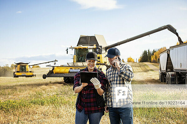 Husband and wife using portable wireless devices to manage and monitor their canola harvest  making a call while combine harvester offloads grain into a semi-trailer grain hauler in the background; Alcomdale  Alberta  Canada