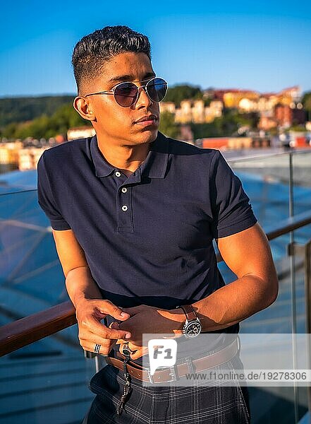 Fashion Lifestyle  Portrait eines jungen Latinos mit der Stadt San Sebastian im Hintergrund  Gipuzkoa. Blaues Polohemd und karierte Hose  nachdenklich auf der Terrasse