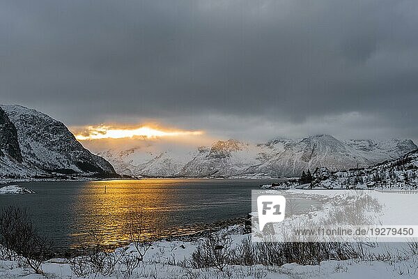 Blick auf die Morgensonne  die durch dichte Wolken hinter schneebedeckten Bergen in der Nähe eines Sees auf den Lofoten in Norwegen bricht