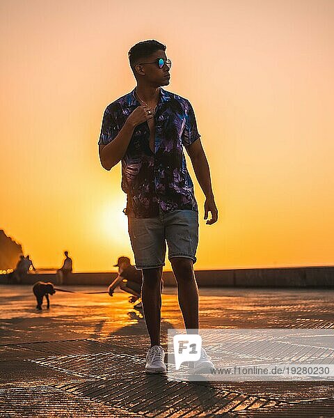 Sommerlicher Lebensstil  ein junger Latino genießt die Sommerferien. mit lila Blumenhemd und Sonnenbrille  schlendert in den Sonnenuntergang