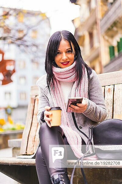 Lifestyle  ein kaukasisches brünettes Mädchen  das mit Papiertüten und einem Kaffee zum Mitnehmen in der Stadt einkauft und dabei eine Nachricht mit dem Telefon verschickt