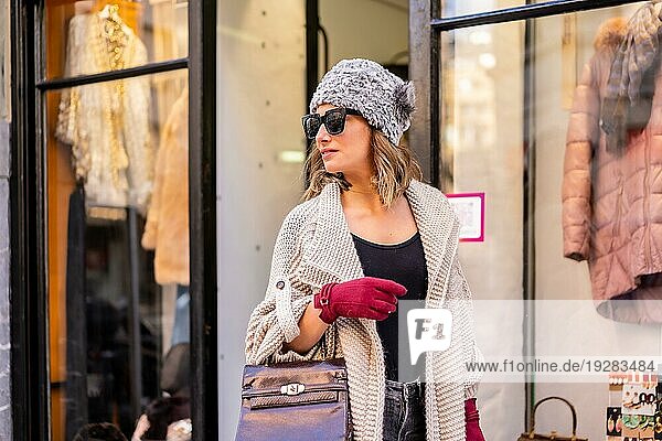 Kaukasisches blondes junges Mädchen genießt das Einkaufen in der Stadt und betrachtet das Schaufenster eines Bekleidungsgeschäfts. Sonnenbrille und rote Handschuhe
