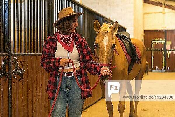 Kaukasische Cowgirl Frau  die sich mit einem Pferd in einem Stall amüsiert und lächelt  amerikanischer Cowboyhut  rotkariertes Hemd und Jeans