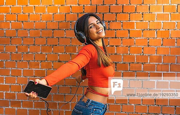 Lifestyle  junge Brünette mit Wollmütze und rotem Pullover mit geschlossenen Augen  die mit ihren schwarzen Kopfhörern Musik von ihrem Handy hört. Hintergrund einer Backsteinmauer