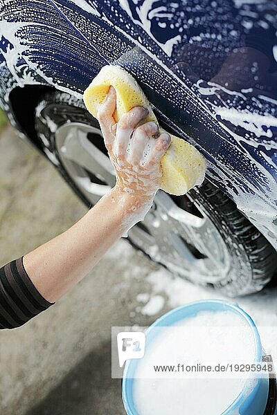 Mann wäscht blaues Auto mit einem gelben Schwamm und einem Eimer Seifenwasser