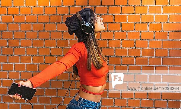 Lifestyle  junge Brünette mit Wollmütze und rotem Pullover mit geschlossenen Augen  die mit ihren schwarzen Kopfhörern Musik von ihrem Handy hört. Hintergrund einer Backsteinmauer