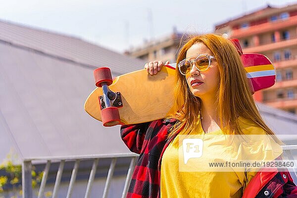 Skateboardfahrerin in gelbem Hemd  rotkariertem Hemd und Sonnenbrille  in einer Pose mit dem Skateboard nach links