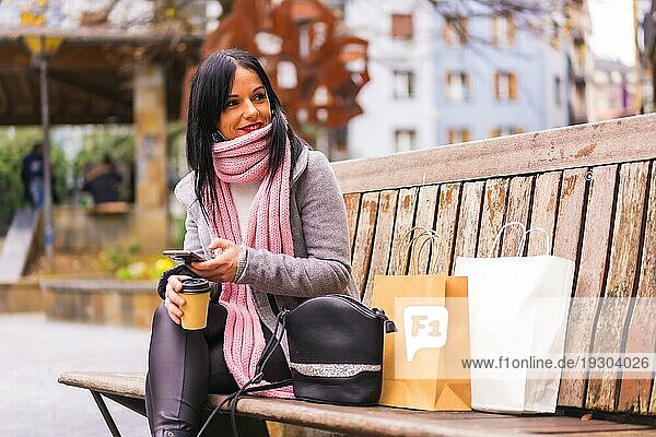 Lifestyle  ein kaukasisches brünettes Mädchen  das mit Papiertüten und einem Kaffee zum Mitnehmen in der Stadt einkauft und dabei eine Nachricht mit dem Telefon verschickt