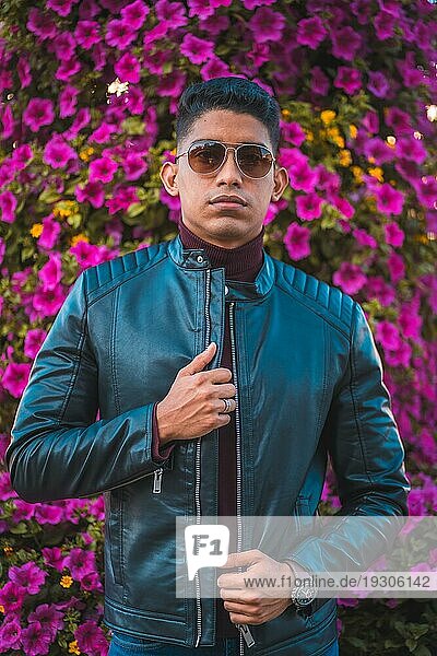 ModeLifestyle Porträt eines jungen Latino mit rosa Blumen im Hintergrund. Jeans  Lederjacke und braune Schuhe