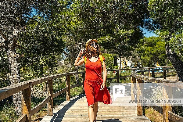 Eine junge Frau in einem roten Kleid geht auf dem Holzsteg in Richtung Strand Moncayo in Guardamar del Segura  Alicante. Gemeinschaft Valencia. Spanien