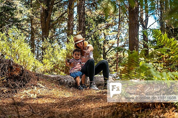 Porträt einer Mutter mit ihrem Sohn  der auf einem Baum in der Natur neben Kiefern sitzt