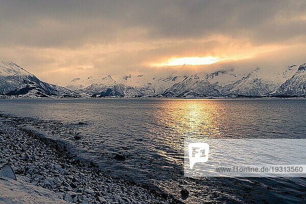 Blick auf die Morgensonne  die durch dichte Wolken hinter schneebedeckten Bergen in der Nähe eines Sees auf den Lofoten in Norwegen bricht