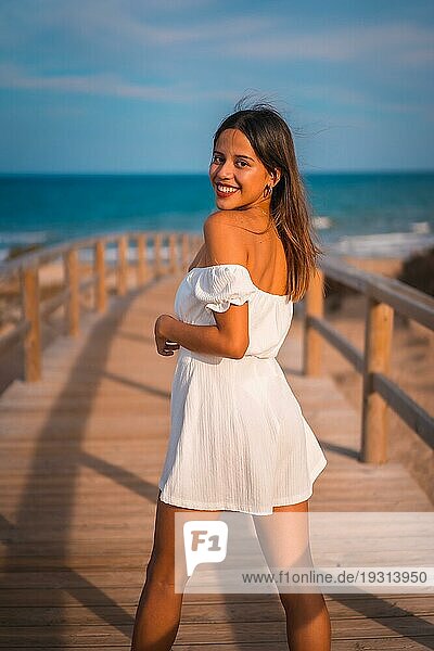 Lebensstil einer jungen brünetten Kaukasierin im Urlaub am Strand  die ein weißes Kleid im Sommer trägt und den Holzsteg entlang geht