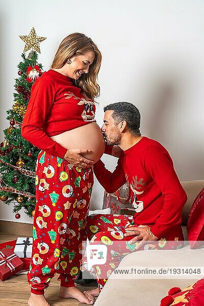 Junges Paar mit Dekoration und roter Weihnachtskleidung küsst den Bauch der schwangeren Freundin. Familie mit schwangerer Frau