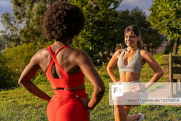 Kaukasisches blondes Mädchen und dunkelhäutiges Mädchen mit Afrohaar machen Stretchingübungen in einem Park. Gesundes Leben  Fitness  Fitness Mädchen  grau und rot Sport Outfits