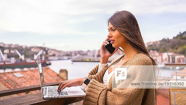 Eine junge lateinamerikanische Frau mit einem weißen TShirt und einem braunen Wollpullover  die auf der Terrasse eines Hauses am Meer telefoniert