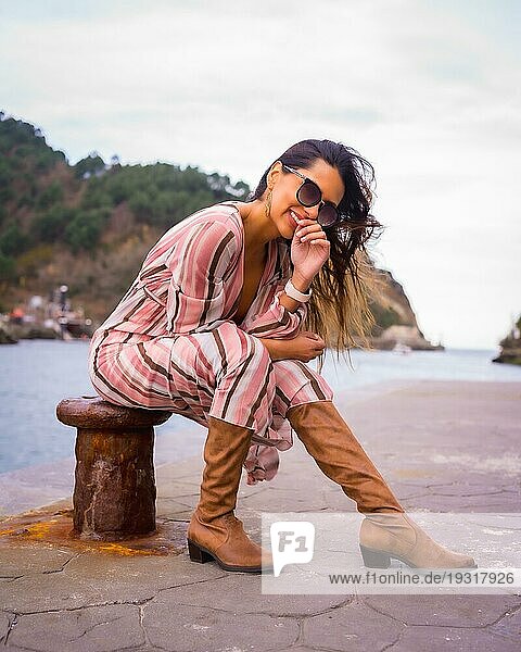 Porträt einer jungen lateinamerikanischen Frau in einem rosa Kleid  braunen Stiefeln und Sonnenbrille  die am Meer sitzend die Sommerferien genießt