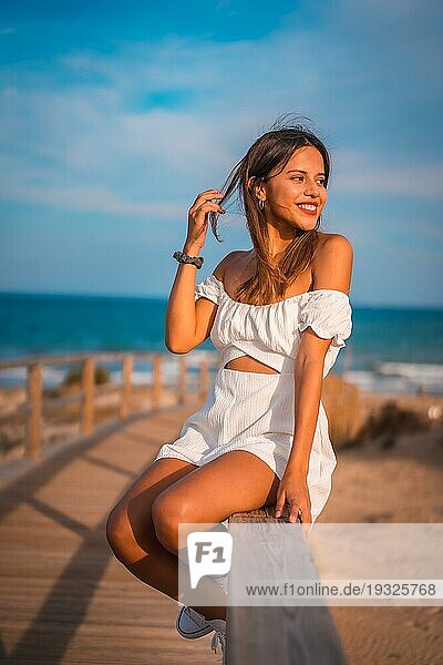 Lebensstil einer jungen brünetten Kaukasierin im Urlaub am Strand in einem weißen Kleid im Sommer  sitzend auf dem Holzsteg