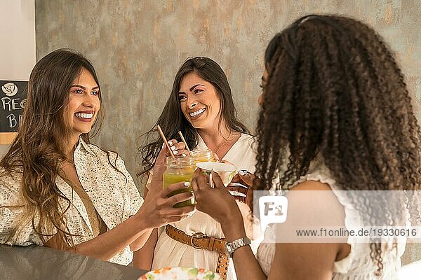 Drei lateinamerikanische glückliche Frauen stoßen mit Säften in einer Cafeteria an
