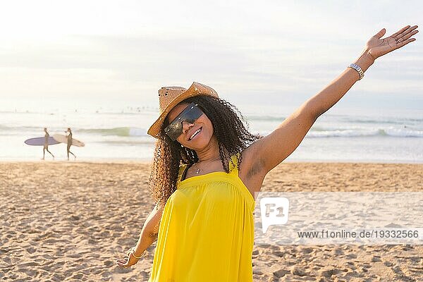 Gemischtrassige glückliche Frau mit Sonnenbrille und Sonnenhut  die die Arme hebt und Wohlbefinden am Strand signalisiert