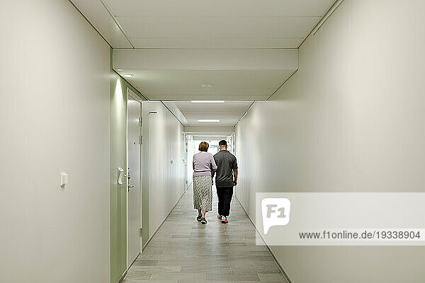 Full length rear view of senior woman walking arm in arm with male caregiver at retirement home corridor