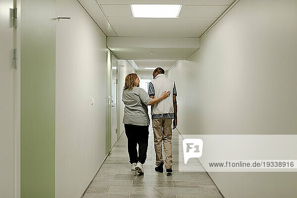 Full length rear view of female nurse walking with arm around senior man in corridor at retirement home