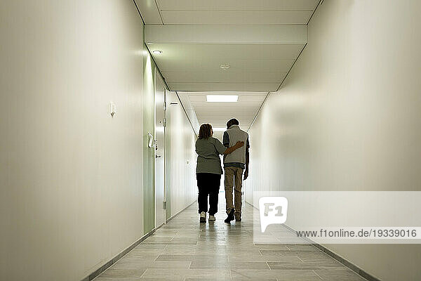 Full length rear view of female caregiver walking with arm around senior man in corridor at nursing home