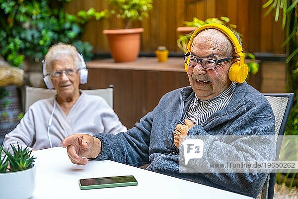 Glücklicher alter Mann  der mit Kopfhörern in einer geriatrischen Einrichtung neben anderen älteren Menschen Musik hört