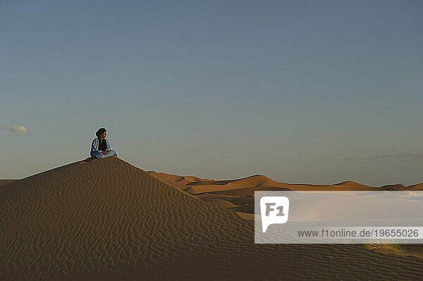 A desert guide in traditional dress sits on a dune at sunset.