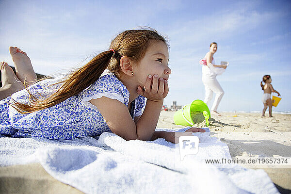 A cute little hispanic girl in a summer dress lays on a towel at the beach.