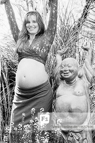 A smiling young pregnant woman standing next to a statue of Budda with her hands arms raised.
