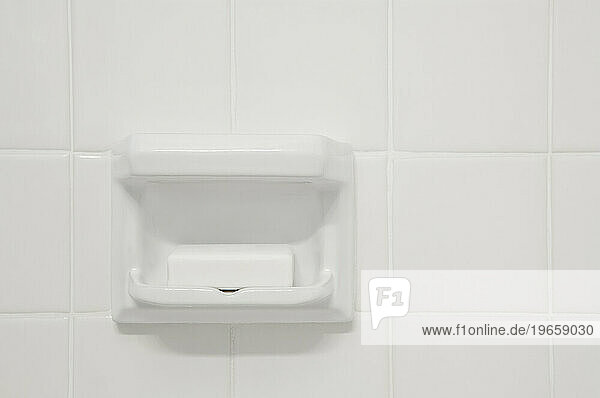 A white tiled wall of a bathroom or shower room with a shaped porcelain recess. A block of soap.