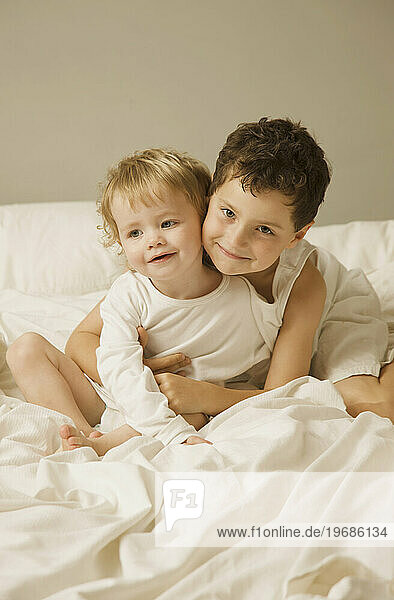 Young boy and toddler sitting in bed hugging and smiling