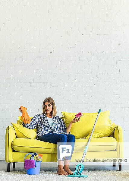 Verwirrte junge Frau sitzt gelbes Sofa hält Handschuhe Pinsel
