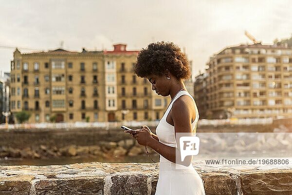 Seitenansicht einer stilvollen afro Frau mit Telefon zu Fuß im Freien während des Sonnenuntergangs neben dem Meer