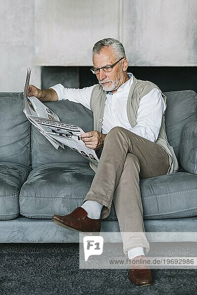 Älterer Mann sitzt gemütlich auf dem Sofa mit gekreuzten Beinen und liest Zeitung