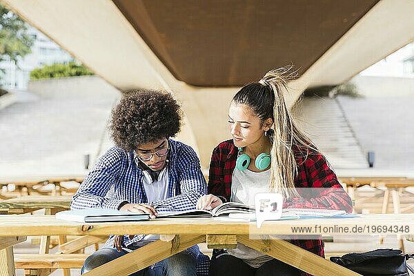 Verschiedenes junges Paar sitzt auf einer Holzbank und studiert zusammen auf dem Universitätscampus