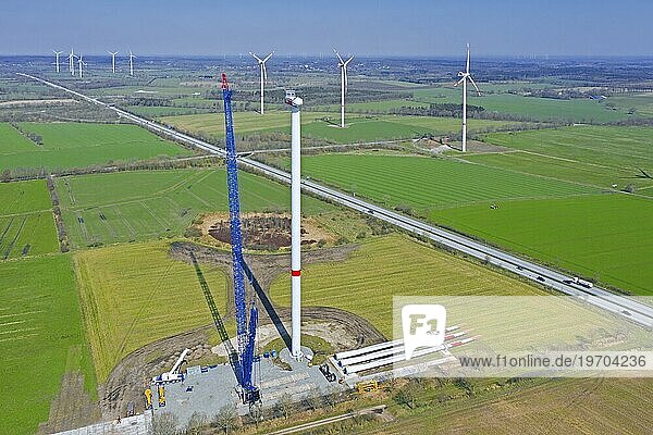 Luftaufnahme der Baustelle mit Rotorblättern am Boden und Baukran  der die Stahlturmteile der Windkraftanlage zusammensetzt