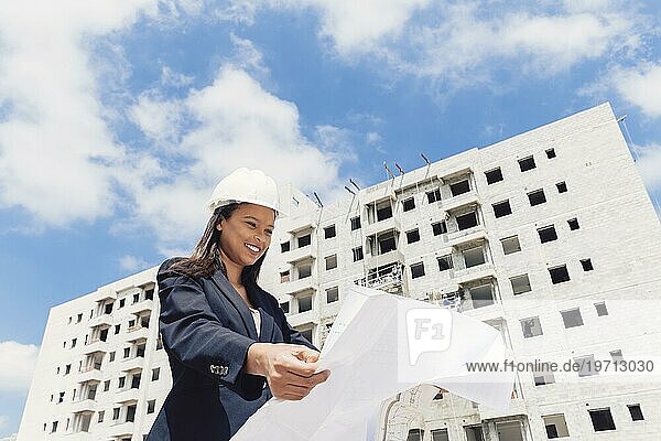 Glückliche afroamerikanische Dame Schutzhelm mit Papier Plan in der Nähe von Gebäudekonstruktion