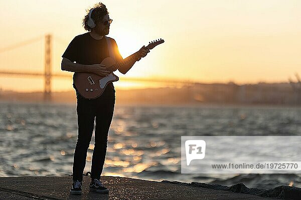 Ein Mann steht am Wasser und spielt Gitarre bei orangefarbenem Sonnenuntergang. Mittlere Einstellung