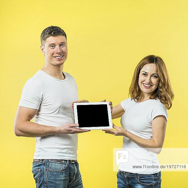 Porträt zwei glückliches junges Paar zeigt schwarzen Bildschirm digitale Tablette gegen gelben Hintergrund
