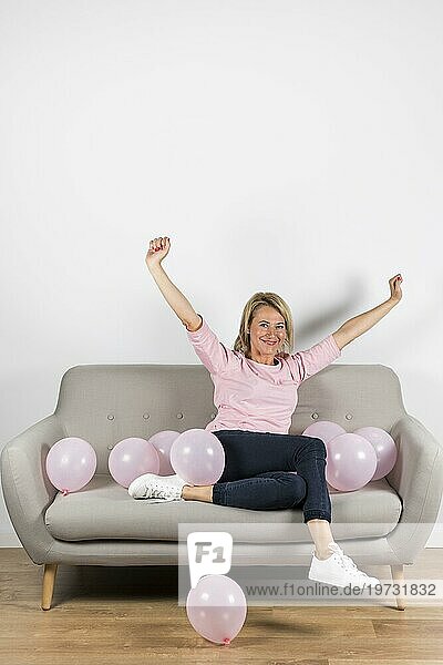 Aufgeregte reife blonde Frau sitzt auf dem Sofa mit rosa Luftballons und hebt ihre Arme
