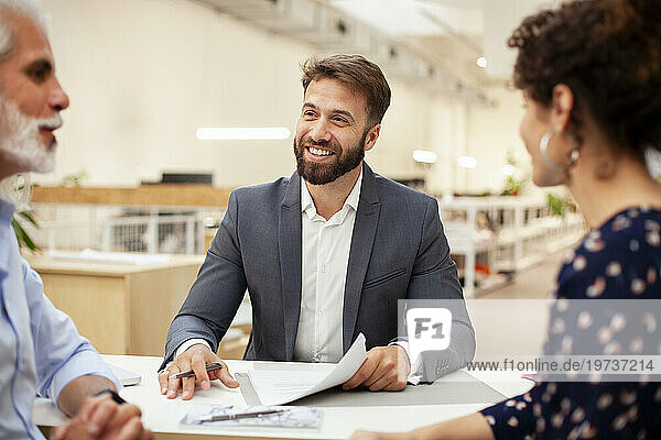 Male real estate agent holding pen and contract while talking to adult couple