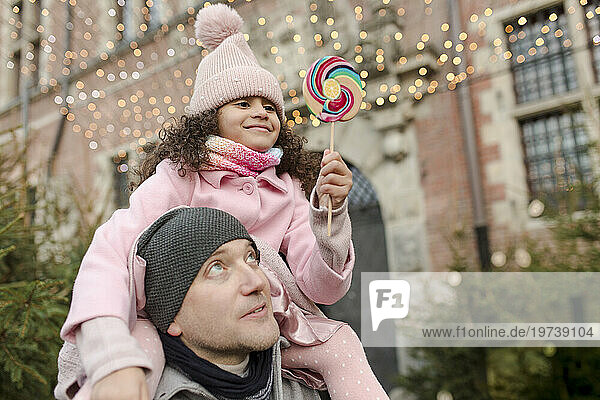 Smiling daughter holding lollipop candy and sitting on father's shoulders