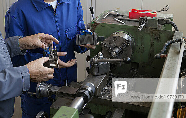 Trainee and instructor holding machine parts near lathe at workshop