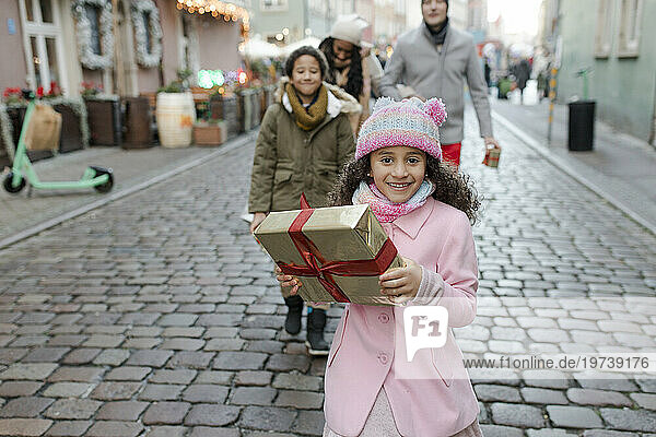 Smiling girl holding gift box and walking with family at Christmas market